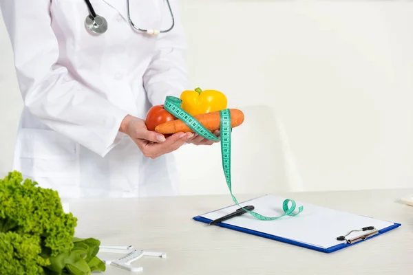 Vista ritagliata di dietista in cappotto bianco che tiene verdure e misura nastro sul posto di lavoro — Foto stock