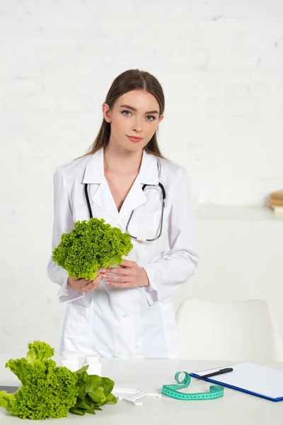 Diätassistentin im weißen Mantel mit Stethocope, das Salat am Arbeitsplatz hält — Stockfoto