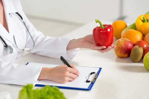 Partial view of dietitian in white coat holding red bell pepper and writing in clipboard at workplace — Stock Photo