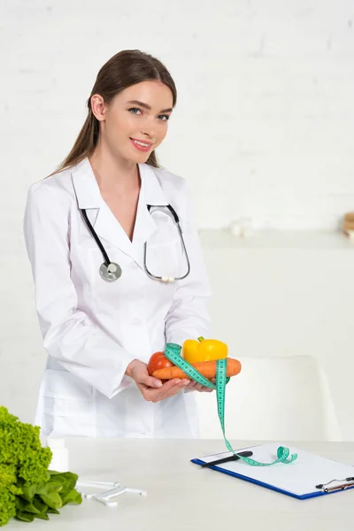 Dietitian in white coat holding vegetables and measure tape at workplace — Stock Photo