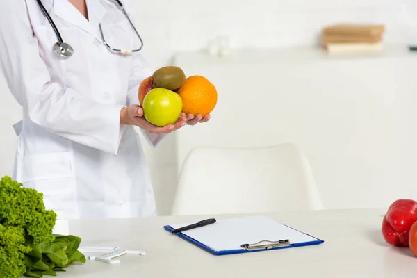 Vista recortada de dietista en bata blanca sosteniendo frutas frescas en el lugar de trabajo - foto de stock