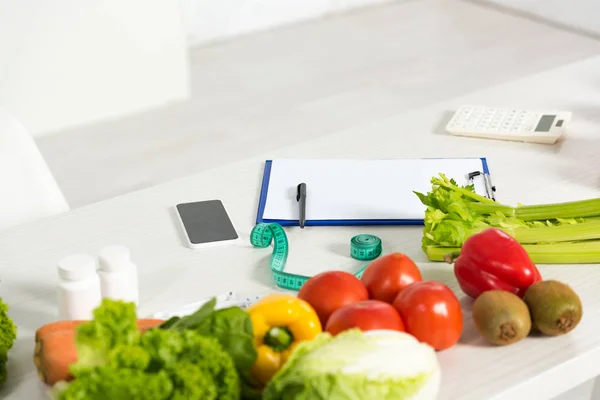 Presse-papiers avec stylo, ruban à mesurer, smartphone avec écran blanc, calculatrice, médicaments et légumes frais sur la table — Photo de stock