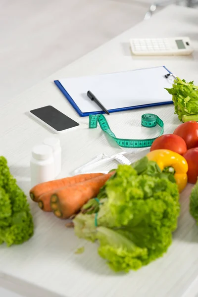 Prancheta com caneta, fita métrica, smartphone com tela em branco, calculadora, remédio e legumes frescos na mesa — Fotografia de Stock