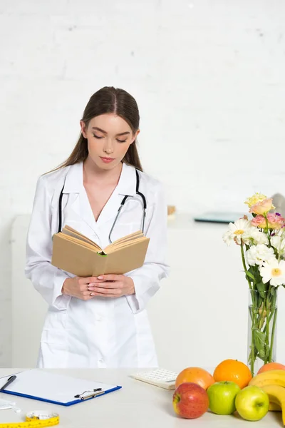 Diététiste concentré en manteau blanc livre de lecture sur le lieu de travail — Photo de stock