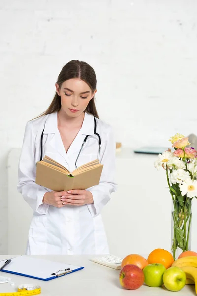 Focused dietitian in white coat reading book at workplace — Stock Photo