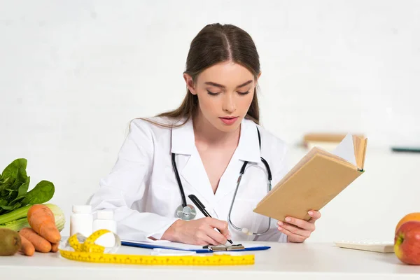 Focused dietitian in white coat reading book and writing in clipboard at workplace — Stock Photo