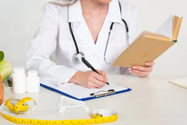 Cropped view of dietitian in white coat reading book and writing in clipboard at workplace — Stock Photo