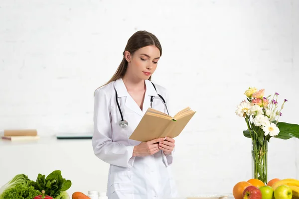 Focused dietitian in white coat reading book at workplace — Stock Photo