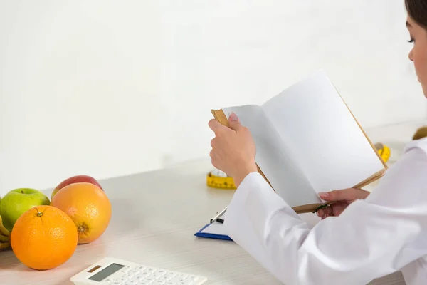 Vista ritagliata di dietista in bianco libro di lettura cappotto sul posto di lavoro — Foto stock