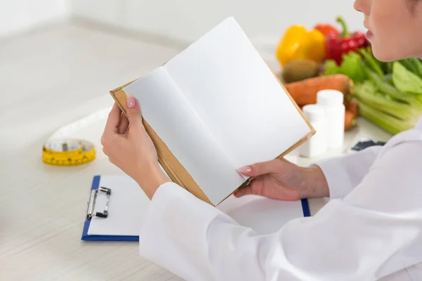 Vista ritagliata di dietista in bianco libro di lettura cappotto sul posto di lavoro — Foto stock