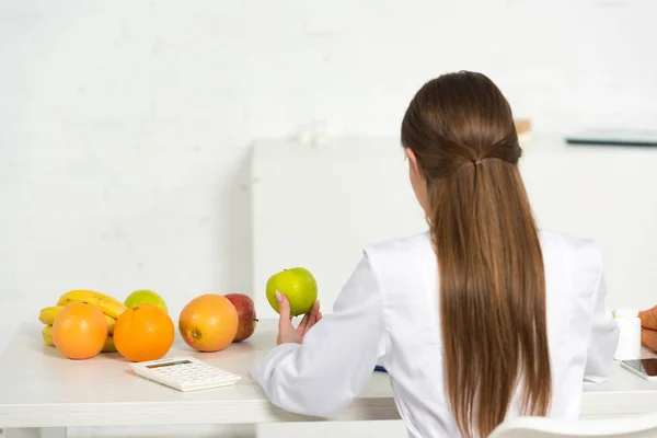 Vista posterior del dietista en bata blanca sosteniendo manzana verde en el lugar de trabajo - foto de stock