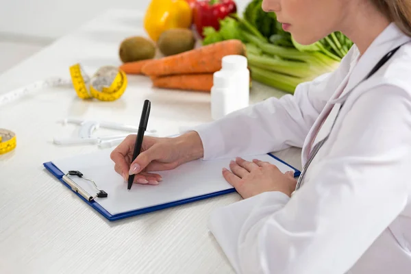 Vista recortada de nutricionista em jaleco branco escrevendo em prancheta no local de trabalho — Fotografia de Stock