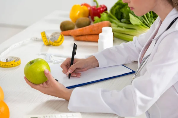 Vue recadrée du diététiste en manteau blanc tenant la pomme verte et écrivant dans le presse-papiers — Photo de stock