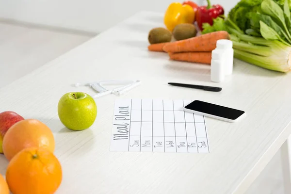 Forfait repas, smartphone avec écran blanc, pilules, stylo, étrier, fruits et légumes frais sur la table — Photo de stock