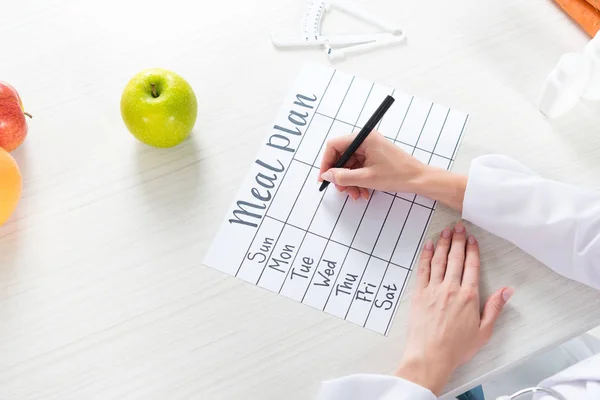 Cropped view of dietitian writing in meal plan at table with fruits — Stock Photo