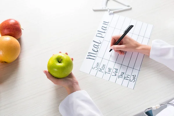Ausgeschnittene Ansicht einer Ernährungsberaterin, die am Arbeitsplatz Apfel in der Hand hält und in den Speiseplan schreibt — Stockfoto