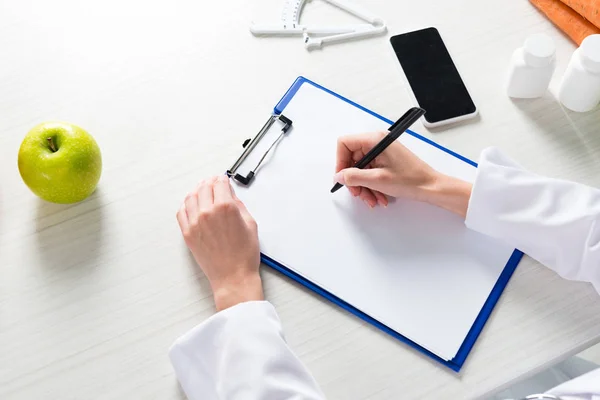 Cropped view of dietitian writing in clipboard at table — Stock Photo