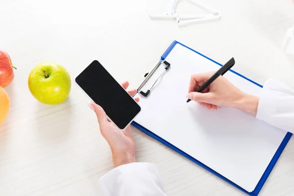 Cropped view of dietitian holding smartphone with blank screen and writing in clipboard — Stock Photo