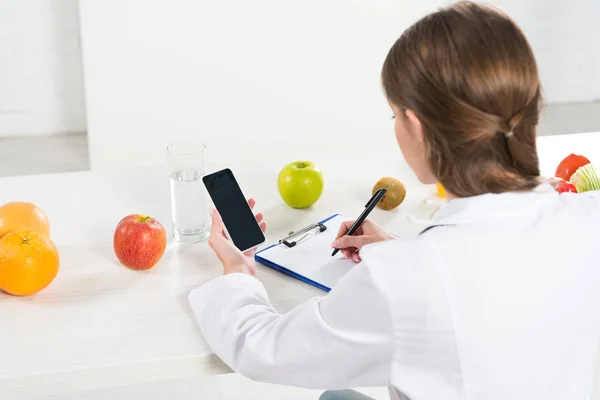 Vista posterior del dietista sosteniendo el teléfono inteligente con pantalla en blanco y escritura en portapapeles en la mesa - foto de stock