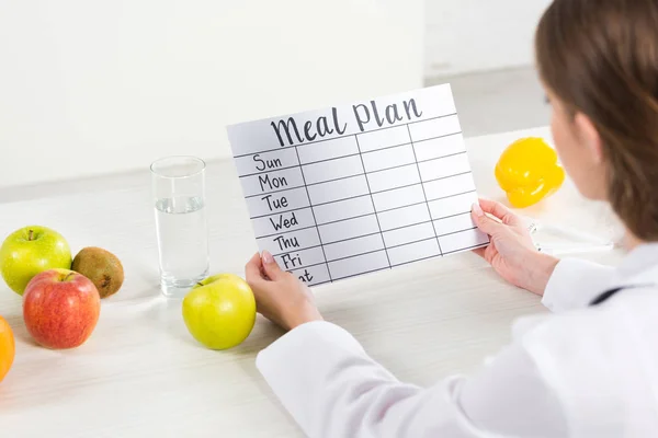 Vista recortada del dietista en bata blanca que sostiene el plan de comidas en el lugar de trabajo - foto de stock