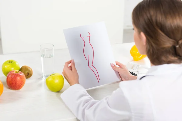 Cropped view of dietitian in white coat holding paper with perfect body image at workplace — Stock Photo