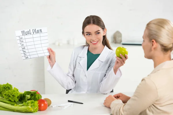 Dietista sonriente en bata blanca sosteniendo plan de comidas y manzana y paciente en la mesa - foto de stock