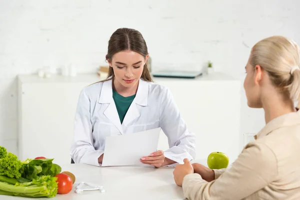 Diététiste en manteau blanc tenant du papier et patient à table — Photo de stock