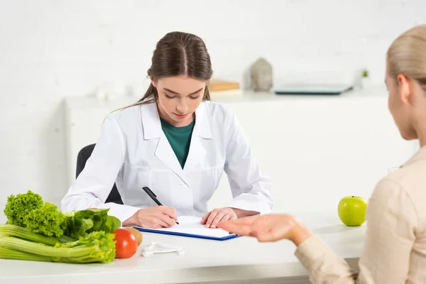 Diététiste concentré en manteau blanc écrit dans le presse-papiers et patient à la table — Photo de stock