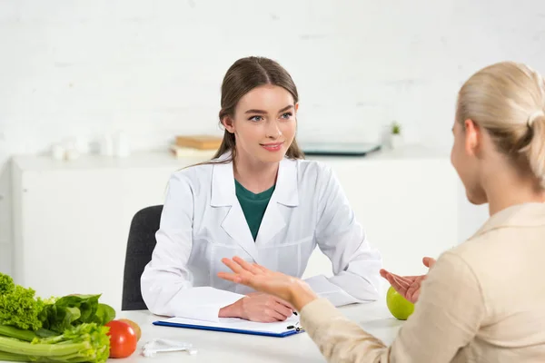 Dietista sonriente en bata blanca mirando al paciente en la mesa - foto de stock