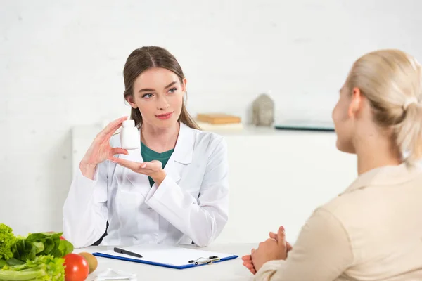 Dietista con bata blanca sosteniendo pastillas y paciente en la mesa - foto de stock