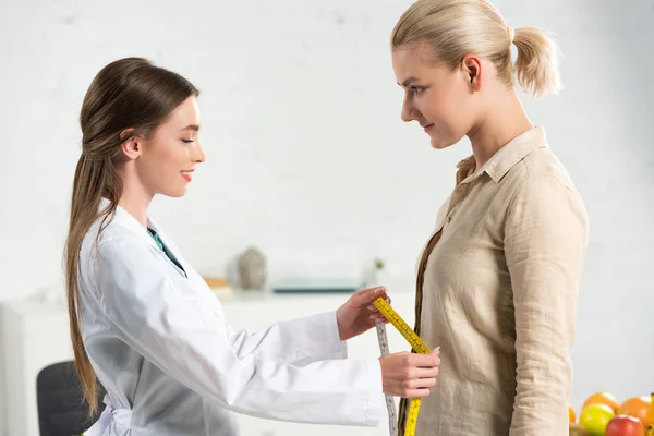 Side view of smiling dietitian in white coat holding measure tape and patient — Stock Photo