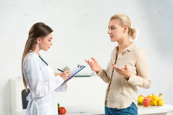 Vue latérale de la diététiste en manteau blanc écrit dans le presse-papiers et le patient — Photo de stock