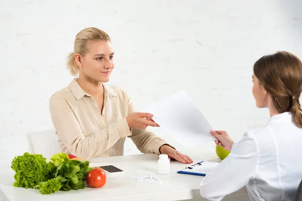 Ernährungsberaterin im weißen Kittel gibt Patientin am Arbeitsplatz Papier — Stockfoto