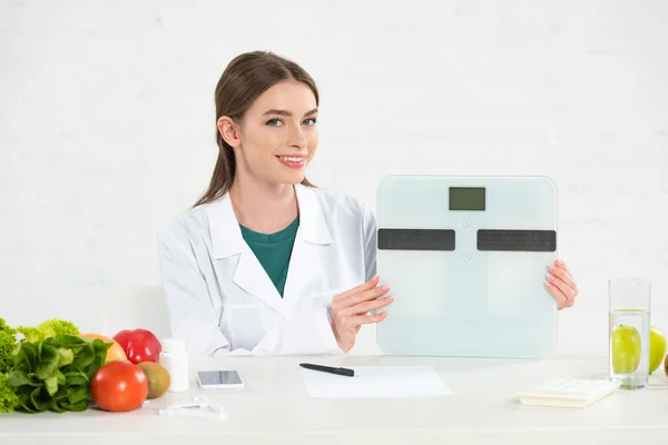 Dietista sonriente en bata blanca sosteniendo escalas digitales en el lugar de trabajo - foto de stock