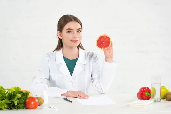Dietista en bata blanca sosteniendo toronja cortada en el lugar de trabajo - foto de stock