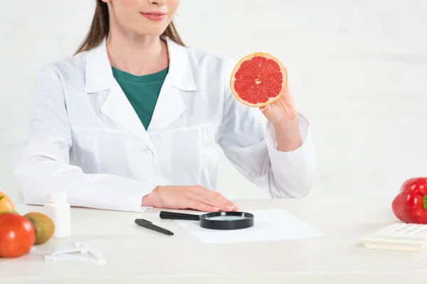 Cropped view of dietitian in white coat holding cut grapefruit at workplace — Stock Photo