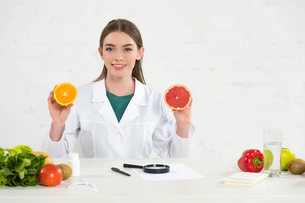 Front view of smiling dietitian in white coat holding cut orange and grapefruit — Stock Photo