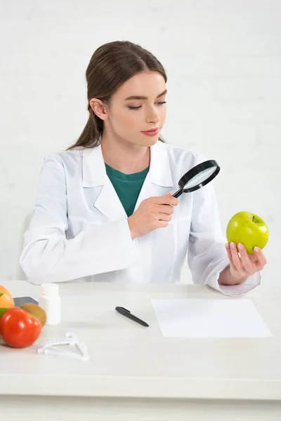 Dietitian in white coat looking at apple through magnifying glass — Stock Photo