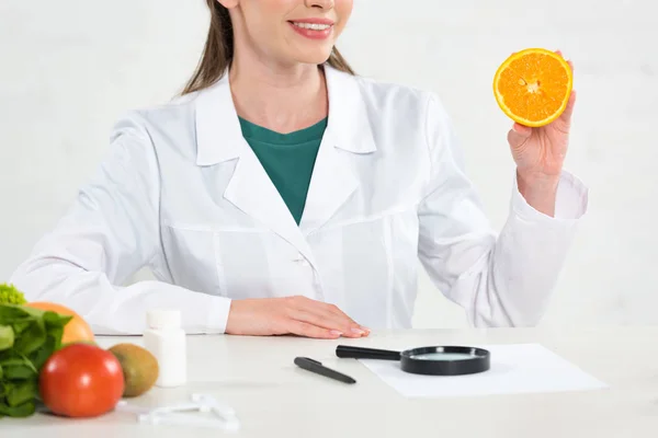 Vista recortada de dietista sonriente en abrigo blanco sosteniendo corte naranja - foto de stock
