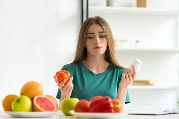 Sad woman sitting at table with fruits and vegetables and holding pills and tomato — Stock Photo