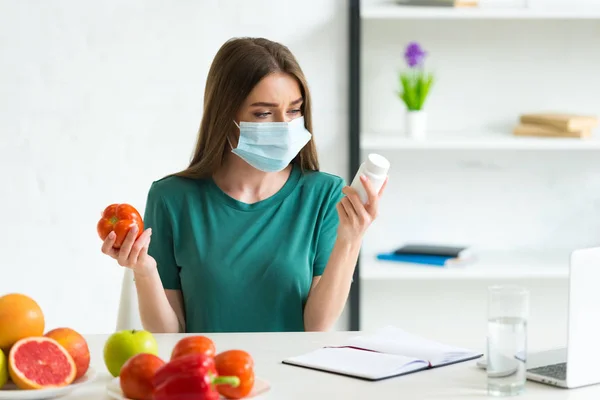 Jovem mulher em máscara médica segurando tomate e pílulas em casa — Fotografia de Stock