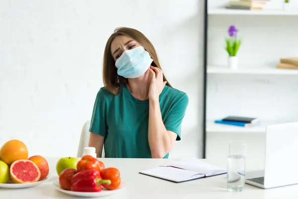 Giovane donna in maschera medica seduta a tavola con frutta, verdura e pillole e graffi al collo a casa — Foto stock