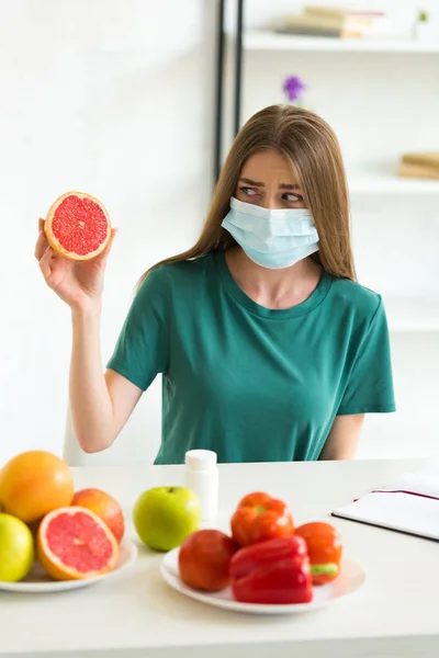 Jovem mulher em máscara médica sentada à mesa com frutas, legumes e pílulas e segurando toranja em casa — Fotografia de Stock