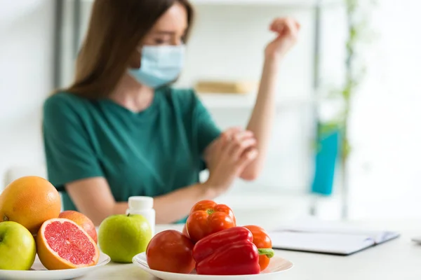 Foco seletivo da jovem mulher em máscara médica sentada à mesa com frutas, legumes e pílulas e coçar o braço em casa — Fotografia de Stock