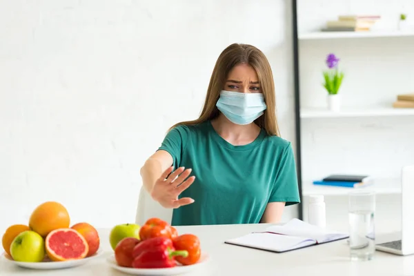 Mujer joven con máscara médica que rechaza frutas y verduras mientras está sentada en la mesa con libro de texto y pastillas en casa - foto de stock