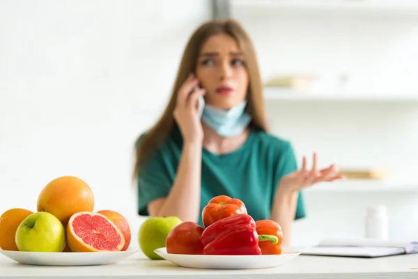 Messa a fuoco selettiva della donna in maschera medica che parla sullo smartphone a tavola con frutta, verdura e pillole a casa — Foto stock
