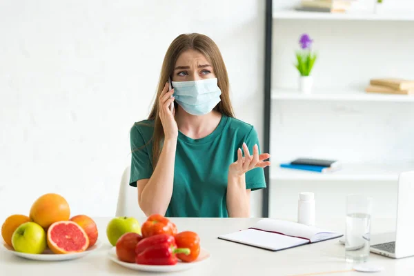 Girl in medical mask talking on smartphone at table with fruits, vegetables and pills at home — Stock Photo