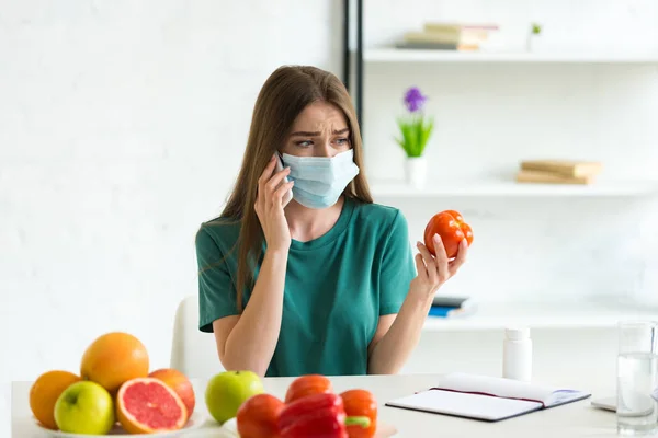 Девушка в медицинской маске разговаривает по смартфону и держит помидор за столом с фруктами, овощами и таблетками дома — стоковое фото