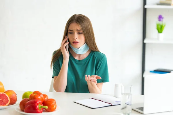 Девушка в медицинской маске разговаривает на смартфоне за столом с фруктами, овощами и таблетками дома — стоковое фото