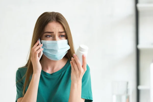 Mujer preocupada en máscara médica hablando en el teléfono inteligente y mirando las píldoras en casa - foto de stock